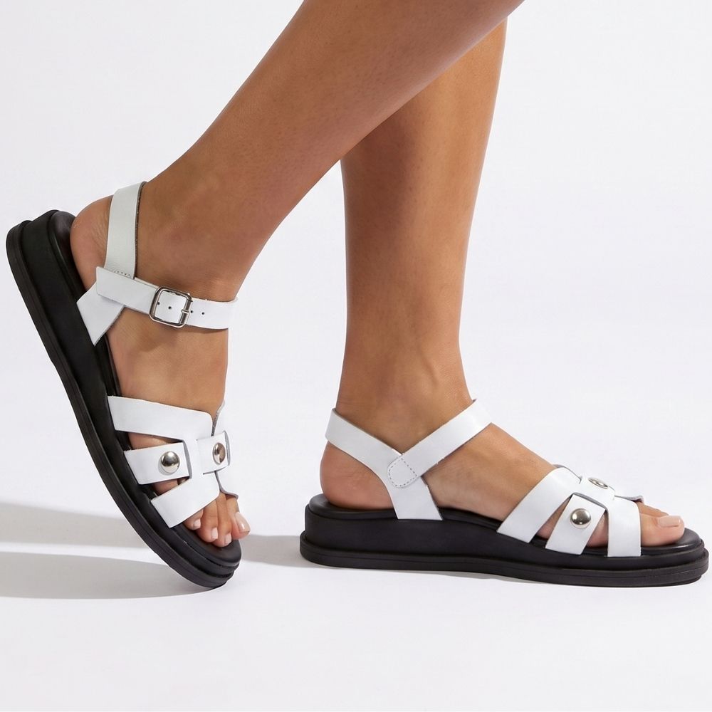 White strappy flat sandals with silver-tone buckle and stud details, shown on a model's feet against a clean white studio background.