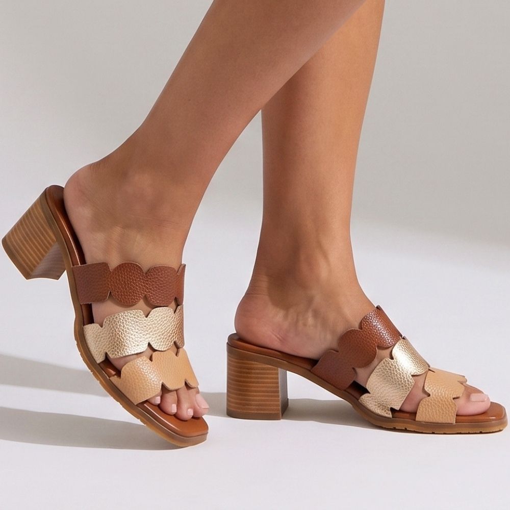 A pair of women's open-toe block-heel mule sandals with geometric straps in tan, beige, and metallic gold, shown on a model's feet against a light gray studio background.