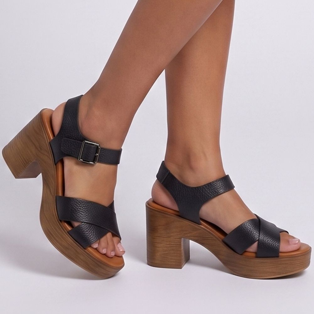 Black leather platform block-heel sandals with crisscross front straps and ankle buckles, shown on a model's feet against a light gray studio background.