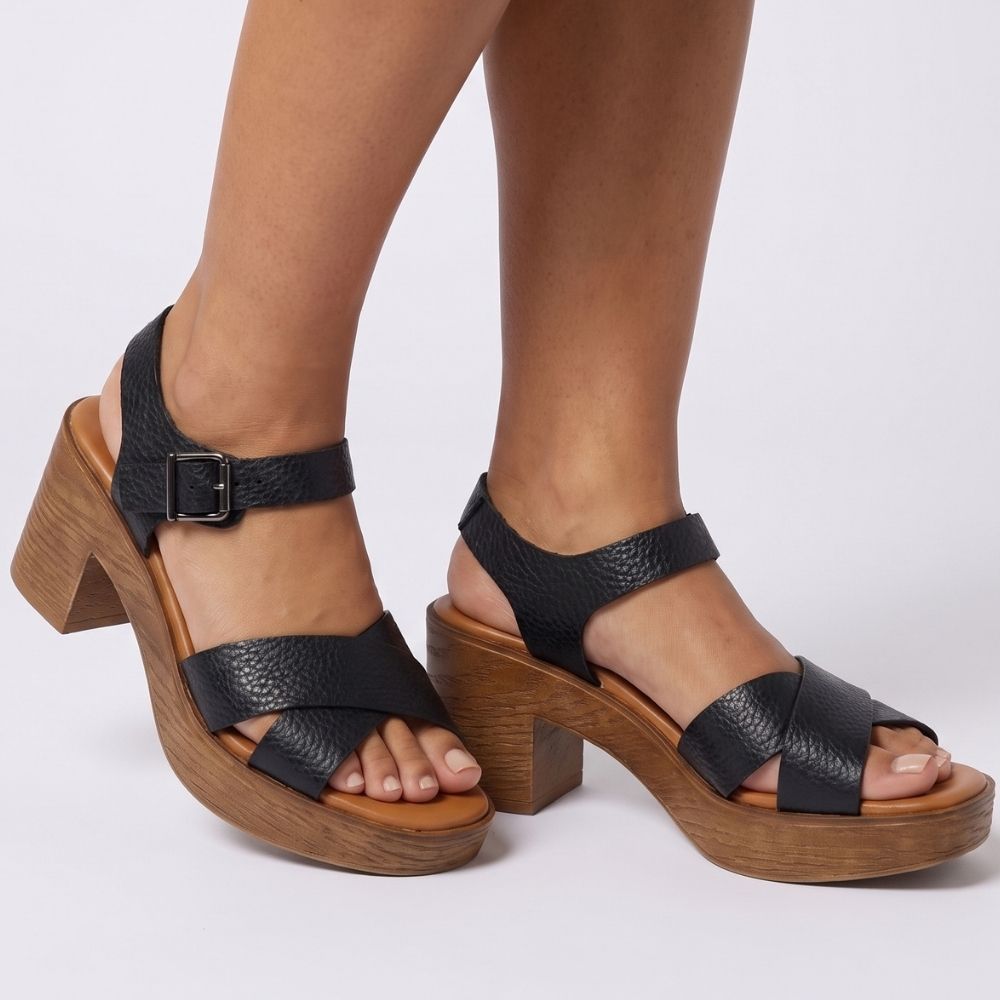 Black pebbled leather platform sandals with wooden block heels and buckled ankle straps, shown on a model's feet against a light gray studio background.