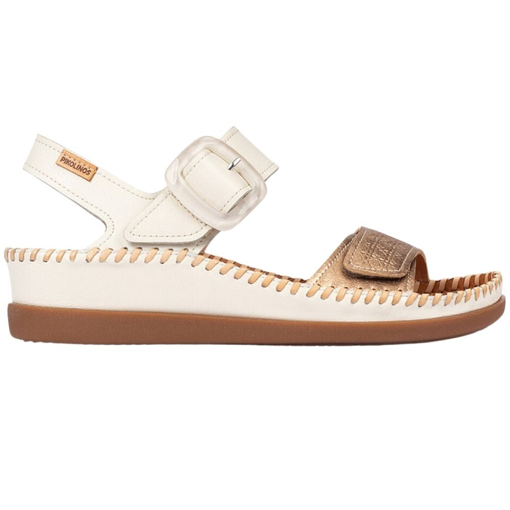 A single sandal from the brand Pikolinos, shown in profile against a white background. The sandal features an off-white leather upper with a decorative stitched edge, a brown sole, a matching off-white ankle strap with a buckle, and a metallic gold toe strap.
