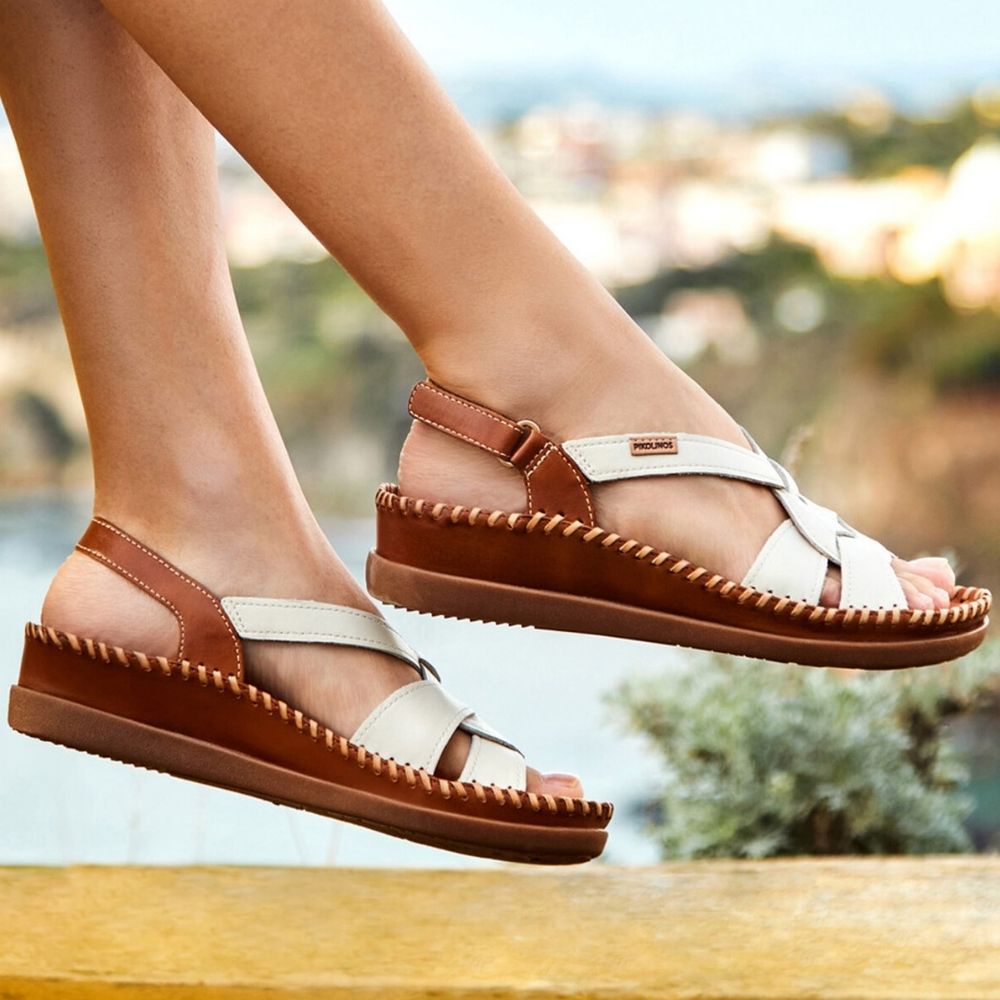 A pair of women's strappy sandals with brown and off-white leather straps, shown being worn on feet against a blurred outdoor background.