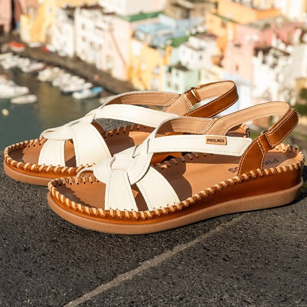 A pair of women's wedge sandals with cream-colored woven straps and tan leather trim, shown on a stone ledge with a blurred coastal town background.