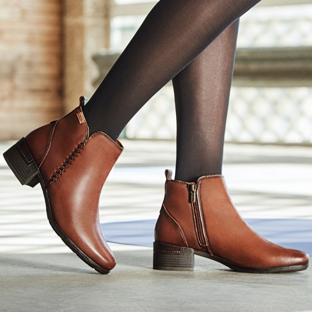 A person wearing brown, leather ankle boots with a low heel and inner zip detailing. The boots have an almond toe and a small, block heel.