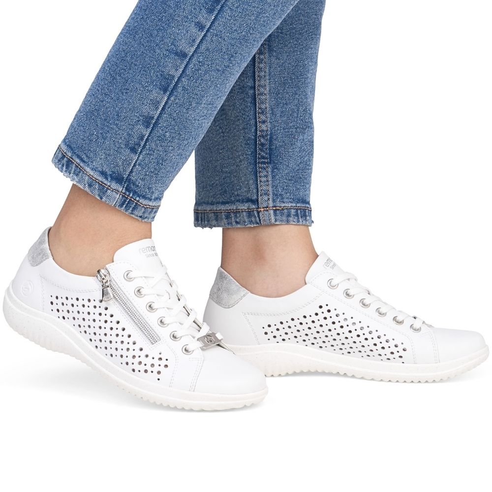 White leather low-top sneakers with perforated side panels, silver zippers, and white laces, shown on a model wearing blue jeans against a white background.