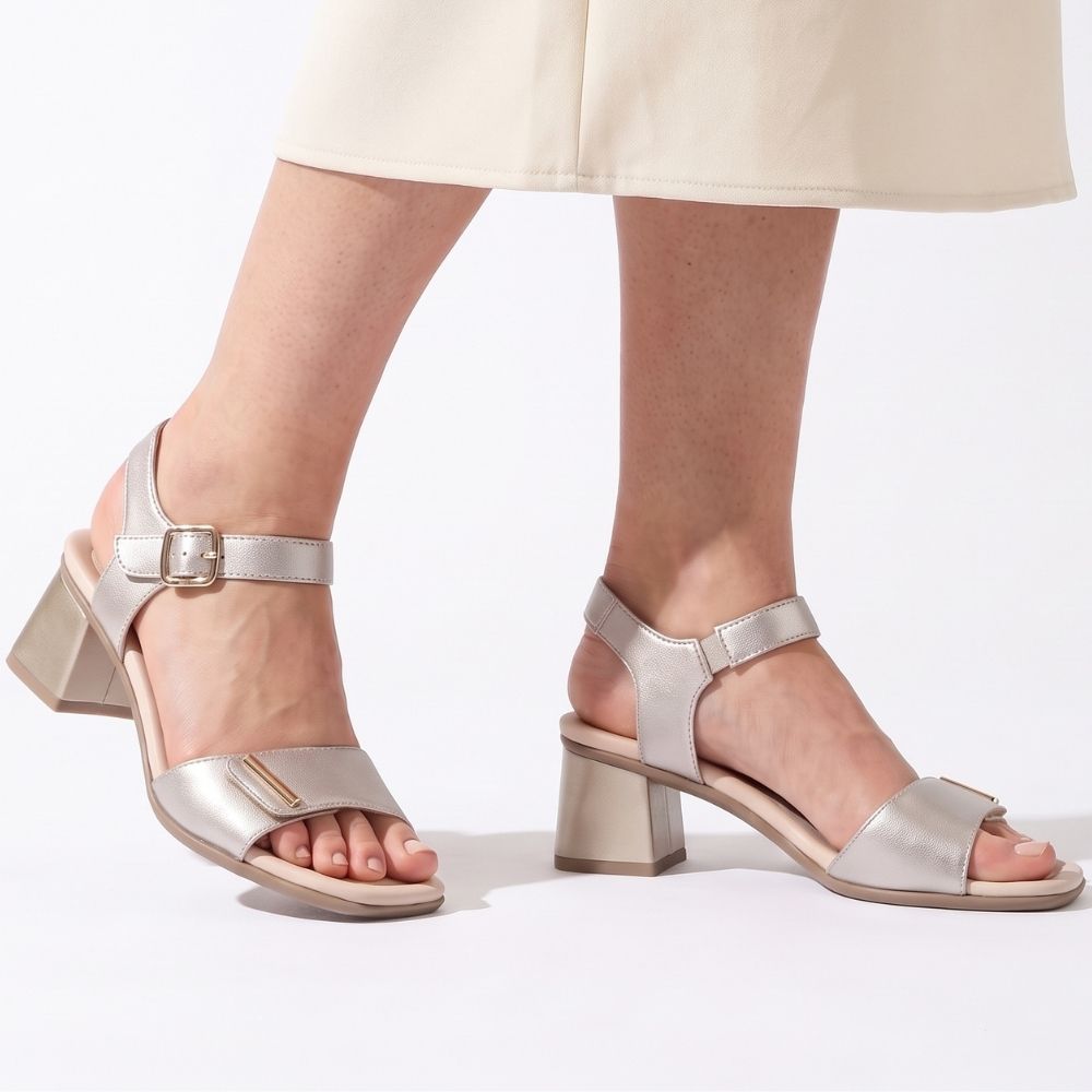 A pair of metallic pale gold block-heel sandals with ankle straps and open toes, shown on a model's feet against a clean white background.