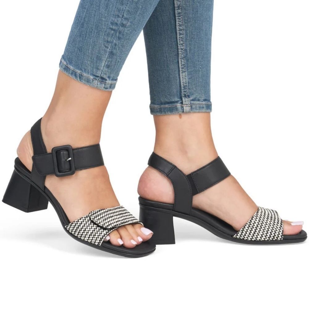 A pair of women's black block-heel sandals with ankle straps and a black-and-beige chevron patterned toe strap, shown on a model's feet wearing blue jeans against a white background.