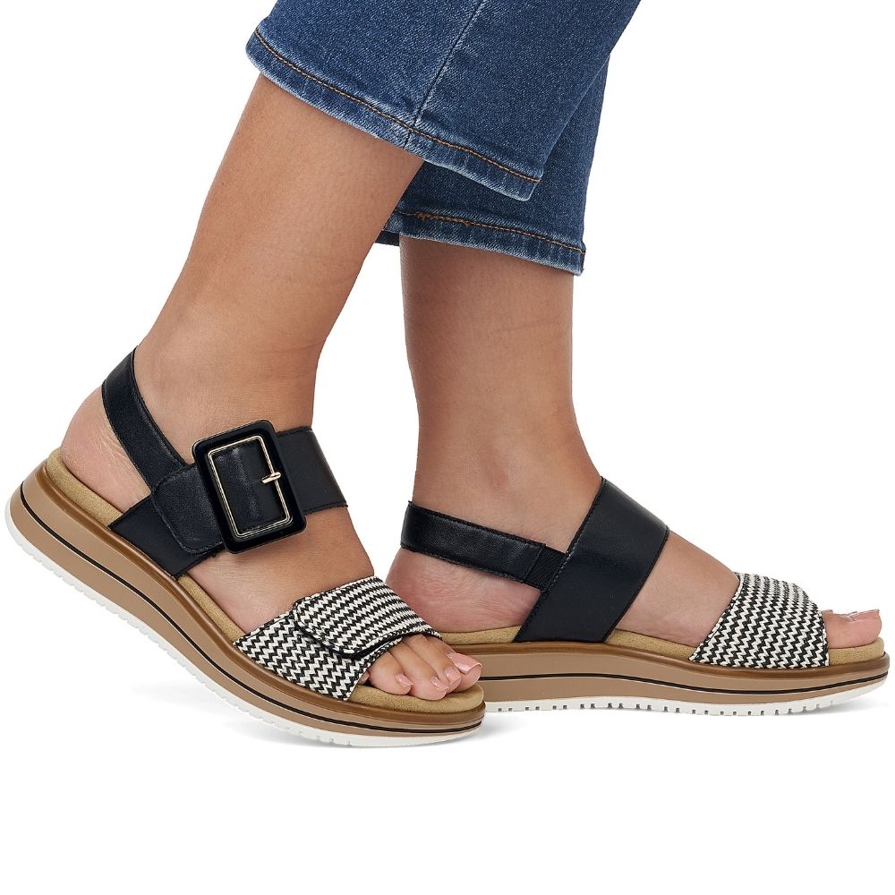 A pair of women's platform sandals shown on a model's feet, featuring black leather straps with a buckle, a black-and-white chevron patterned toe strap, and a tan platform sole.