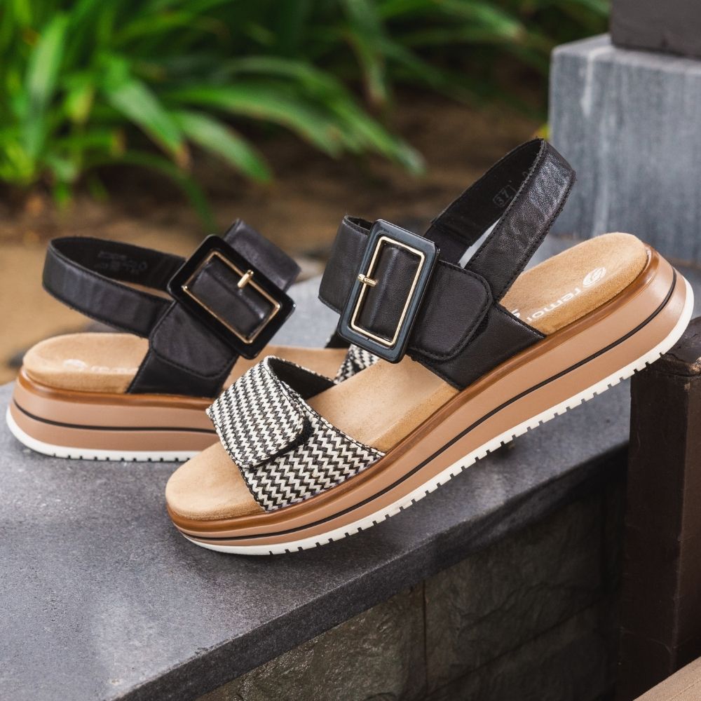 A pair of women's platform sandals with black leather straps, gold-tone buckles, and a black-and-white chevron pattern across the front, resting on a dark stone ledge with green foliage in the background.