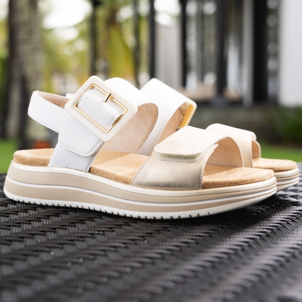 A pair of women's platform sandals with white and pale gold leather straps, a gold buckle, and a beige textured footbed, resting on a black woven outdoor chair with a blurred garden background.