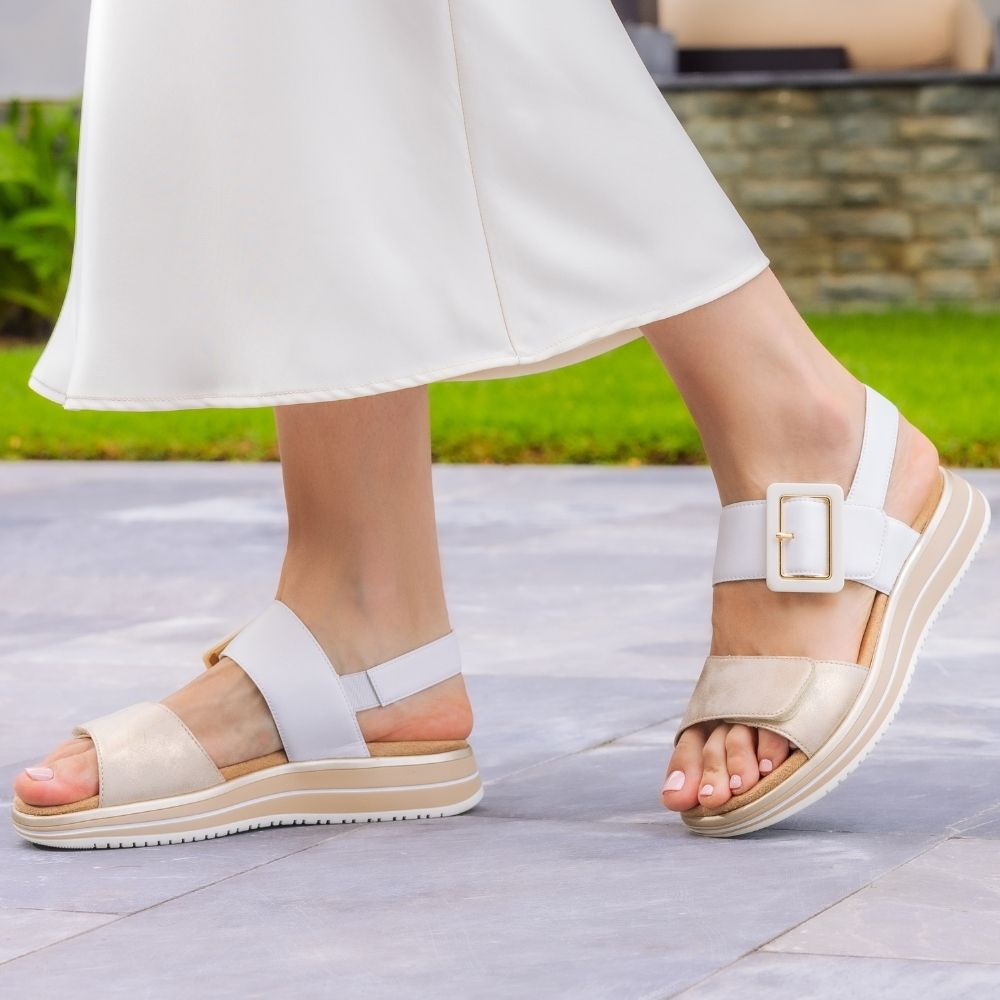 A pair of women's platform sandals shown on a model's feet, featuring a wide pale gold toe strap, a white ankle strap with a square buckle, and a chunky beige and white platform sole, set against a blurred outdoor patio background.