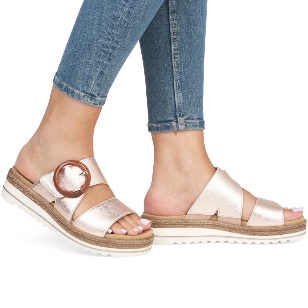 A pair of women's open-toe platform slide sandals in a metallic rose gold leather, featuring a statement buckle on the side strap, worn with blue jeans against a white background.