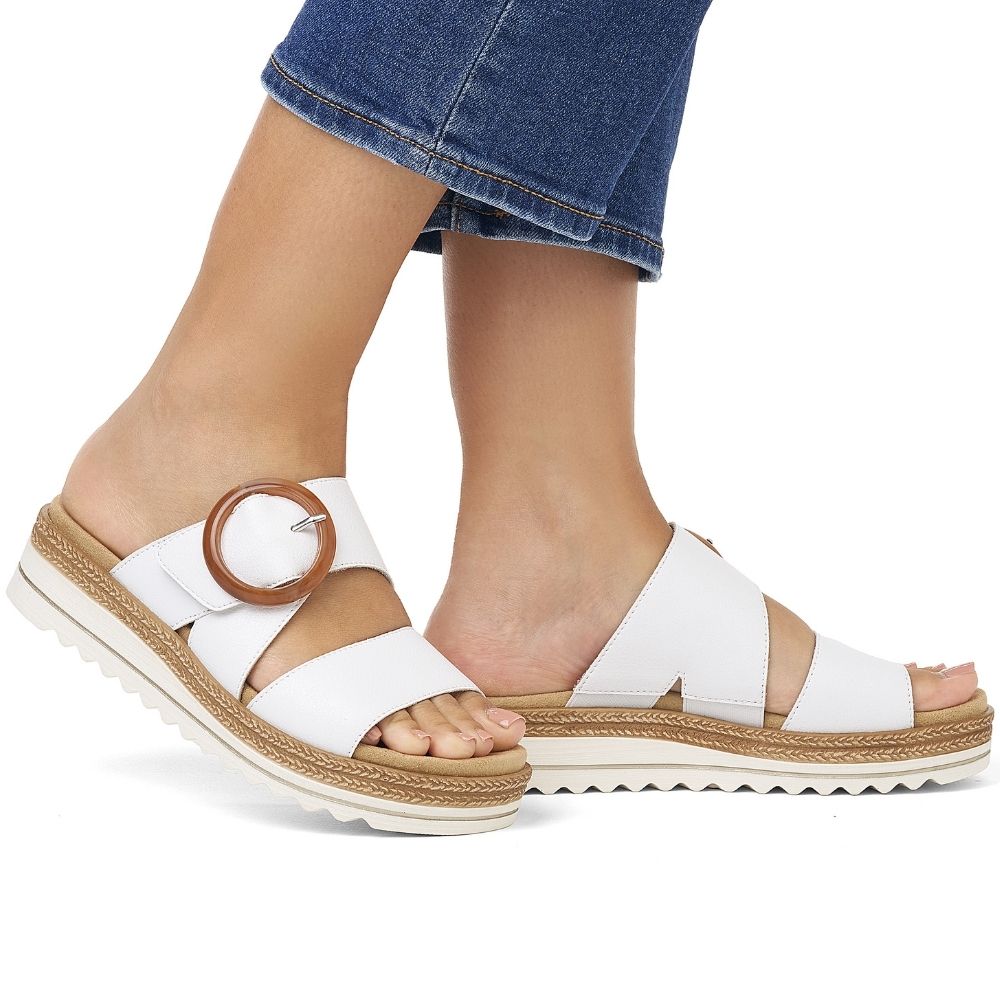 White leather slide sandals with a statement amber circular buckle on the strap and a white sole with a braided jute-look trim, shown on a model's feet with blue jeans against a white background.