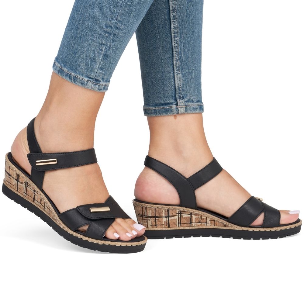 Black leather wedge sandals with ankle and toe straps with gold accents, shown on a model wearing blue jeans against a white background.