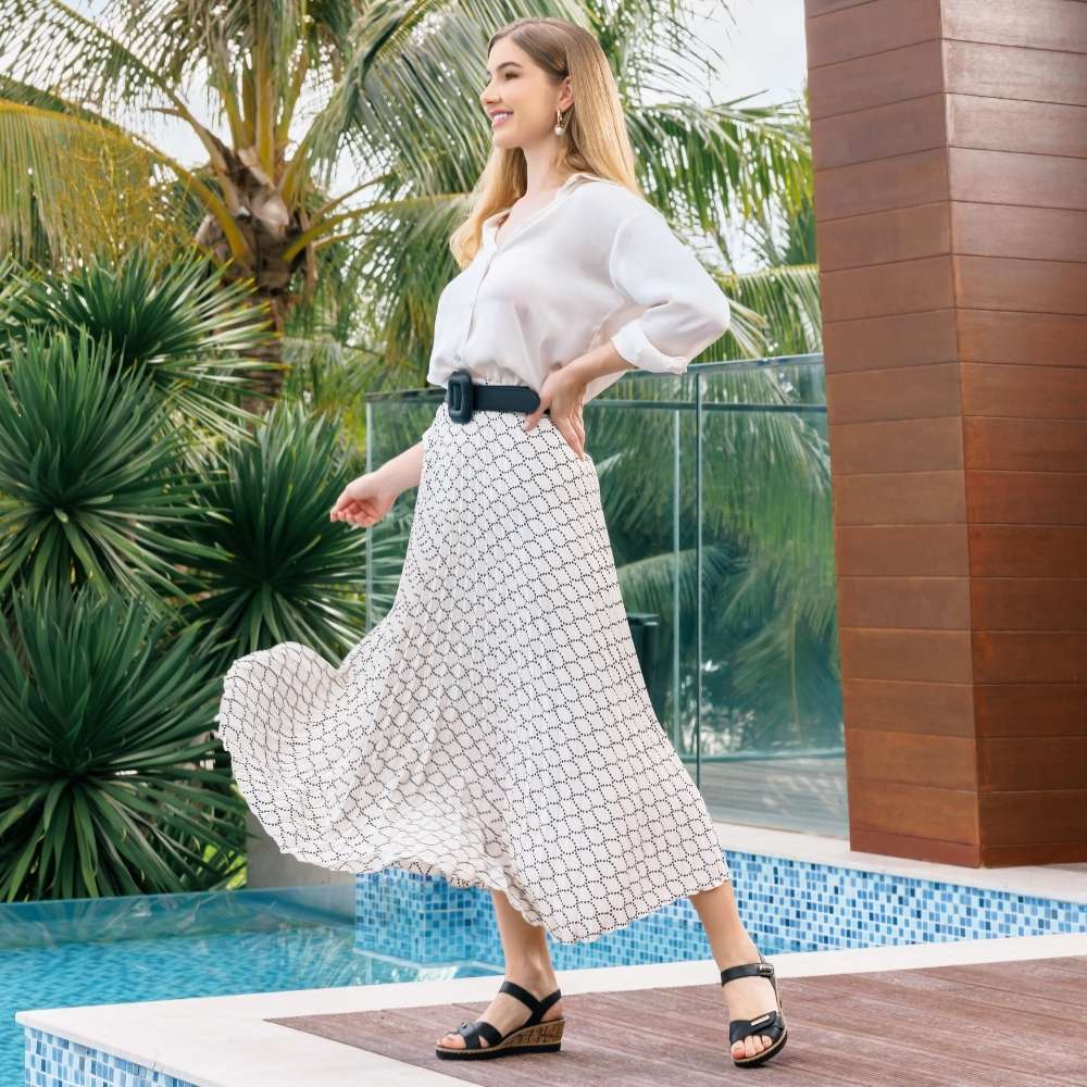 A woman stands by a resort pool wearing a white long-sleeve blouse and a white maxi skirt with a black geometric pattern, paired with a black belt and black wedge sandals.