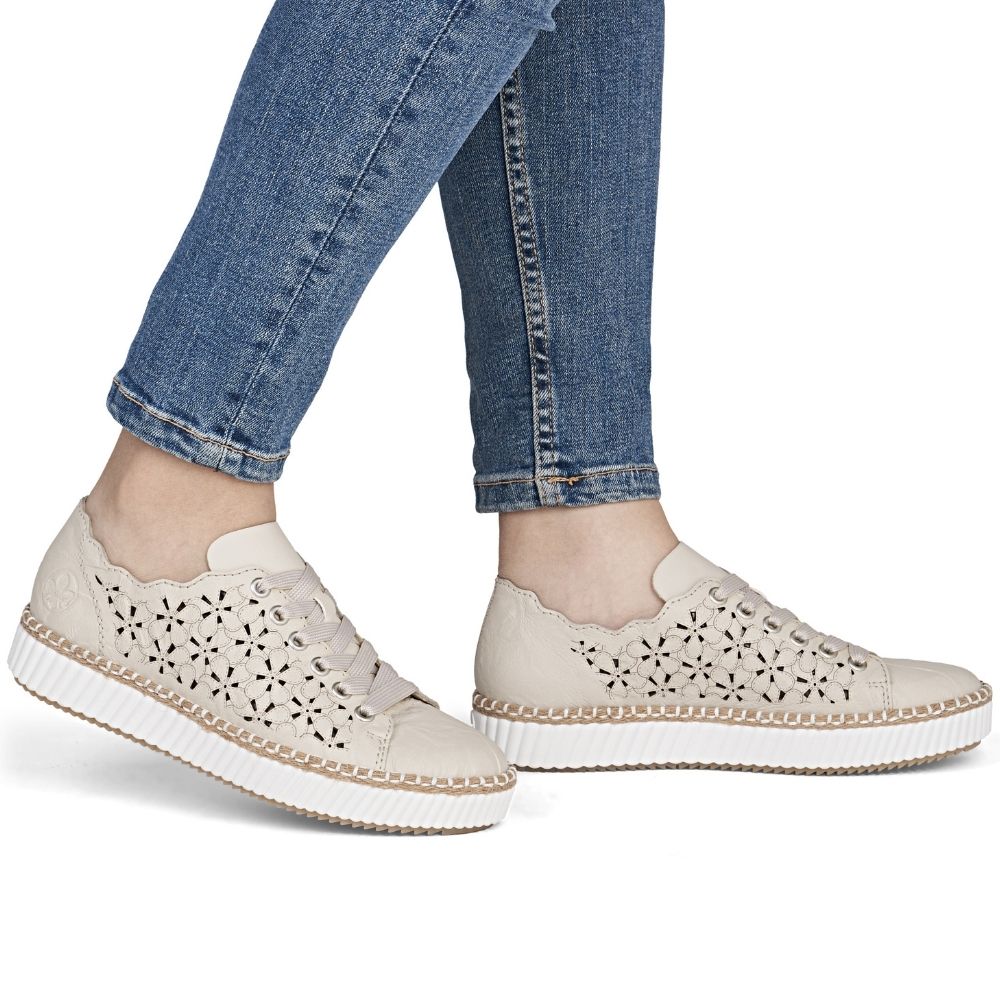Cream perforated lace-up sneakers with a thick white sole, shown on a model wearing blue jeans against a white background.