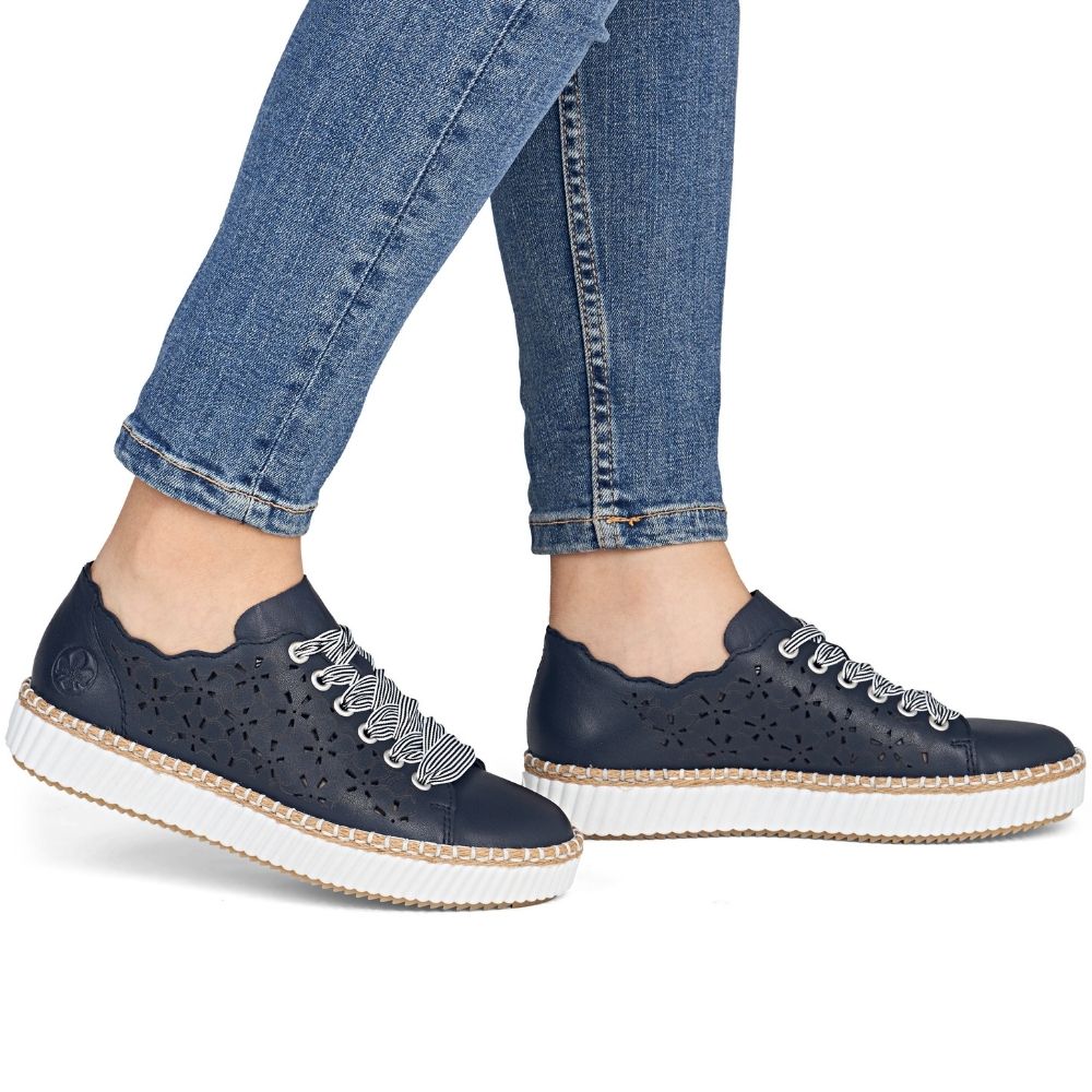 A pair of navy-blue leather platform sneakers with scalloped edges, perforated detailing, and white laces, shown on a model wearing blue jeans against a white background.