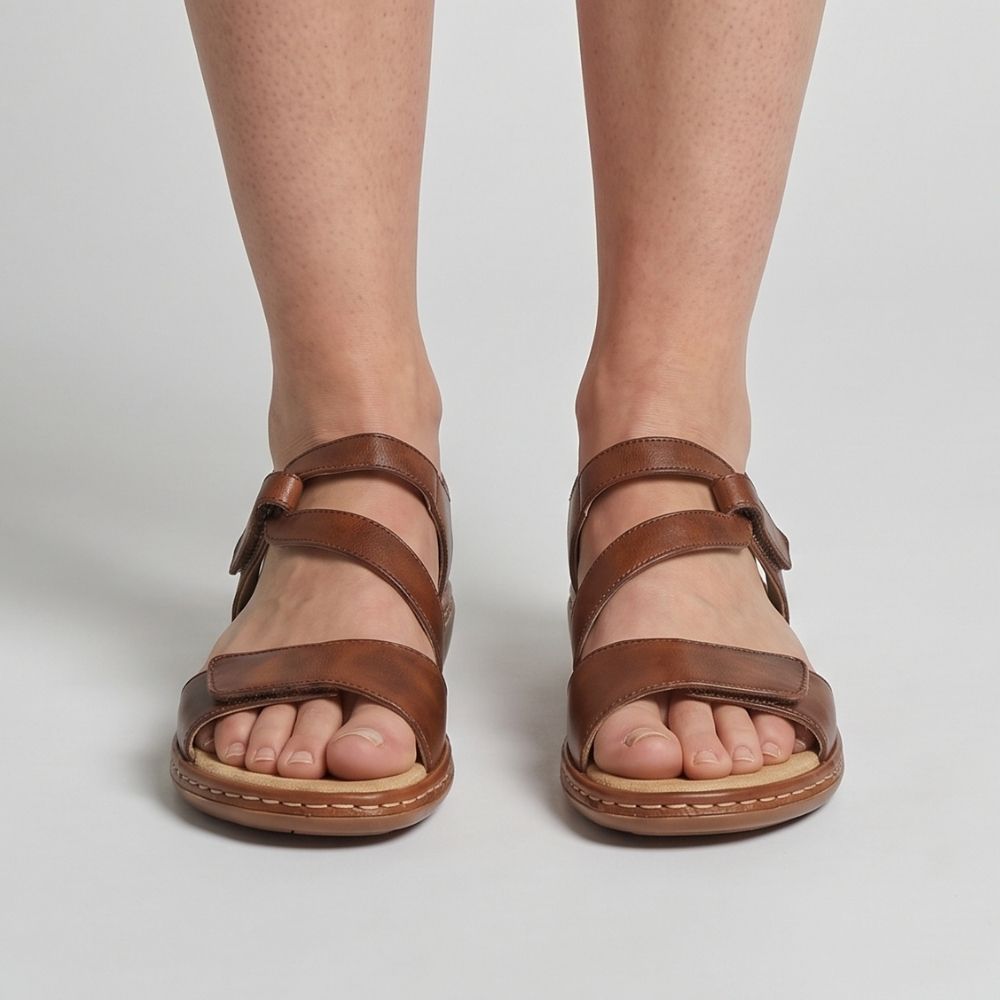 A pair of women's tan leather strappy sandals with a low wedge heel, shown on a model's feet against a light gray studio background.