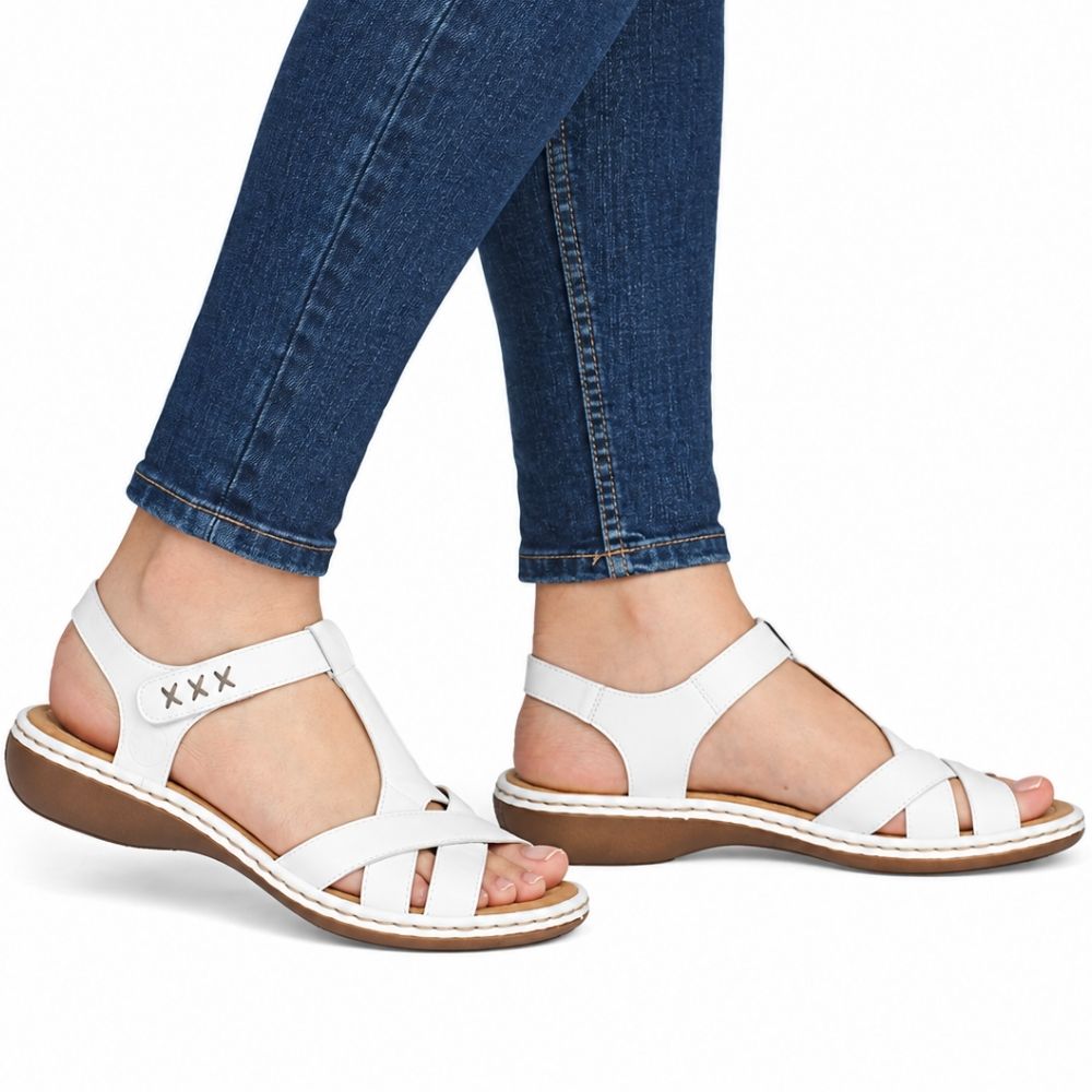 White leather T-strap sandals with a low wedge heel, shown on a model's feet wearing blue skinny jeans against a white background.