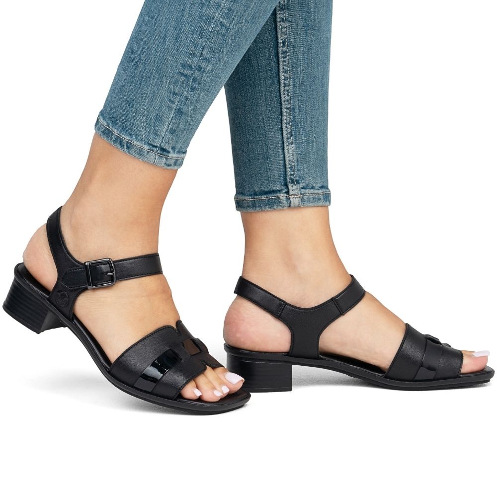 A pair of black leather block-heel sandals with cutout straps, shown on a model's feet paired with cuffed blue jeans against a white background.