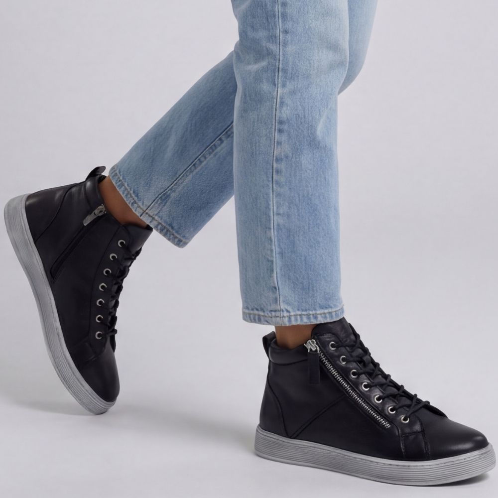 Black high-top sneaker boots with zippers worn with light blue jeans on a gray background