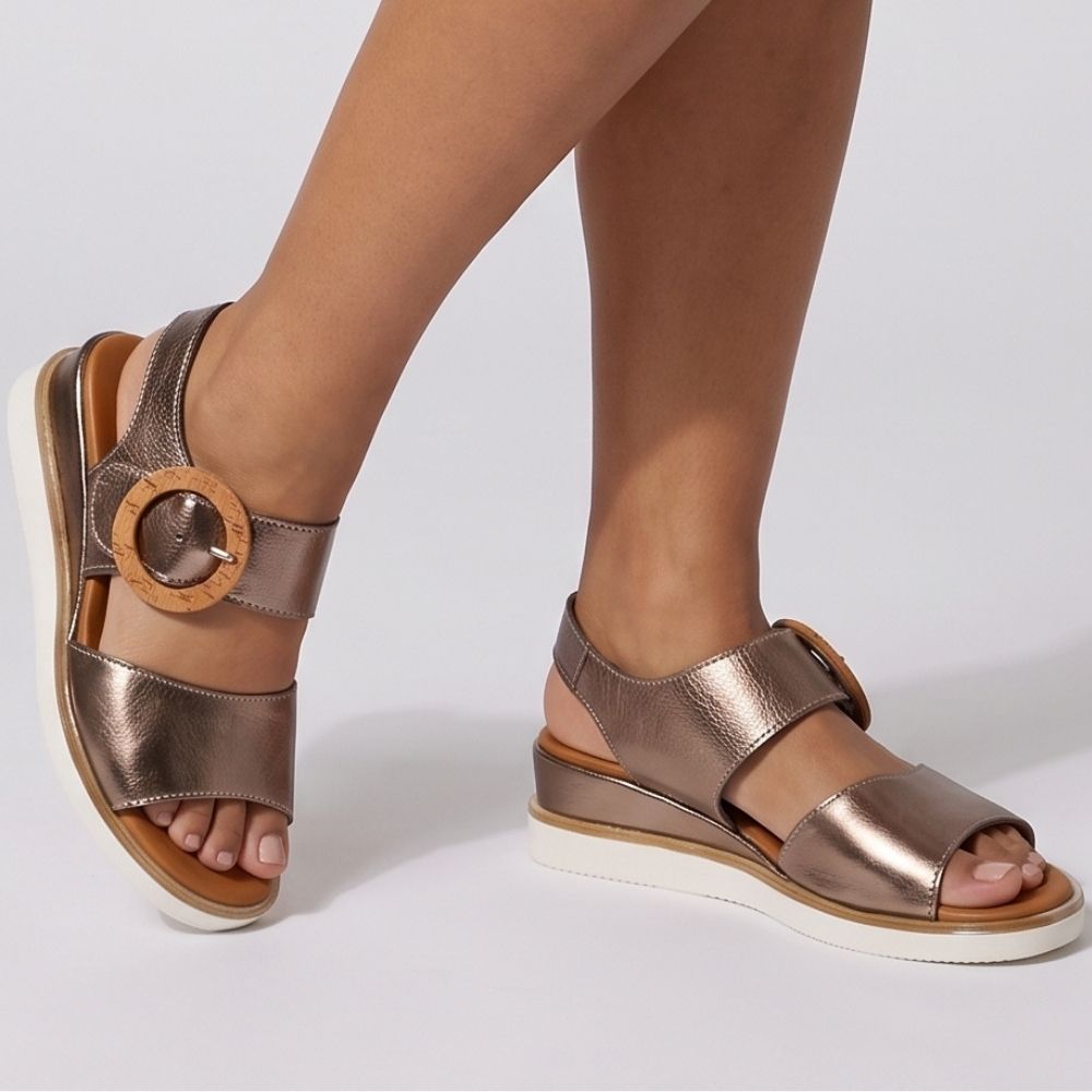 Metallic bronze leather wedge sandals with circular wooden buckles, shown on a model's feet against a light gray studio background.