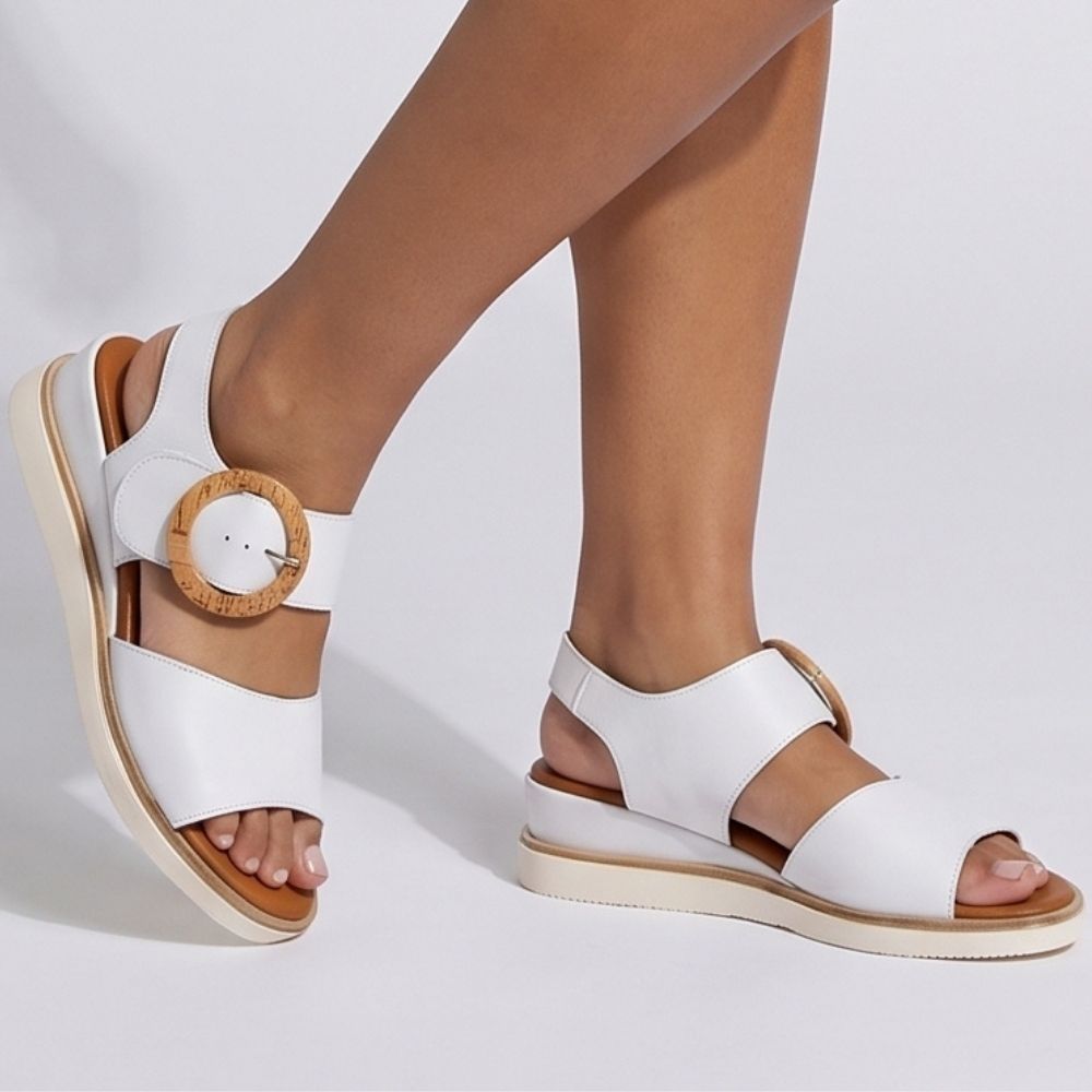 White leather wedge sandals with circular wooden buckles, shown on a model's feet against a light gray studio background.