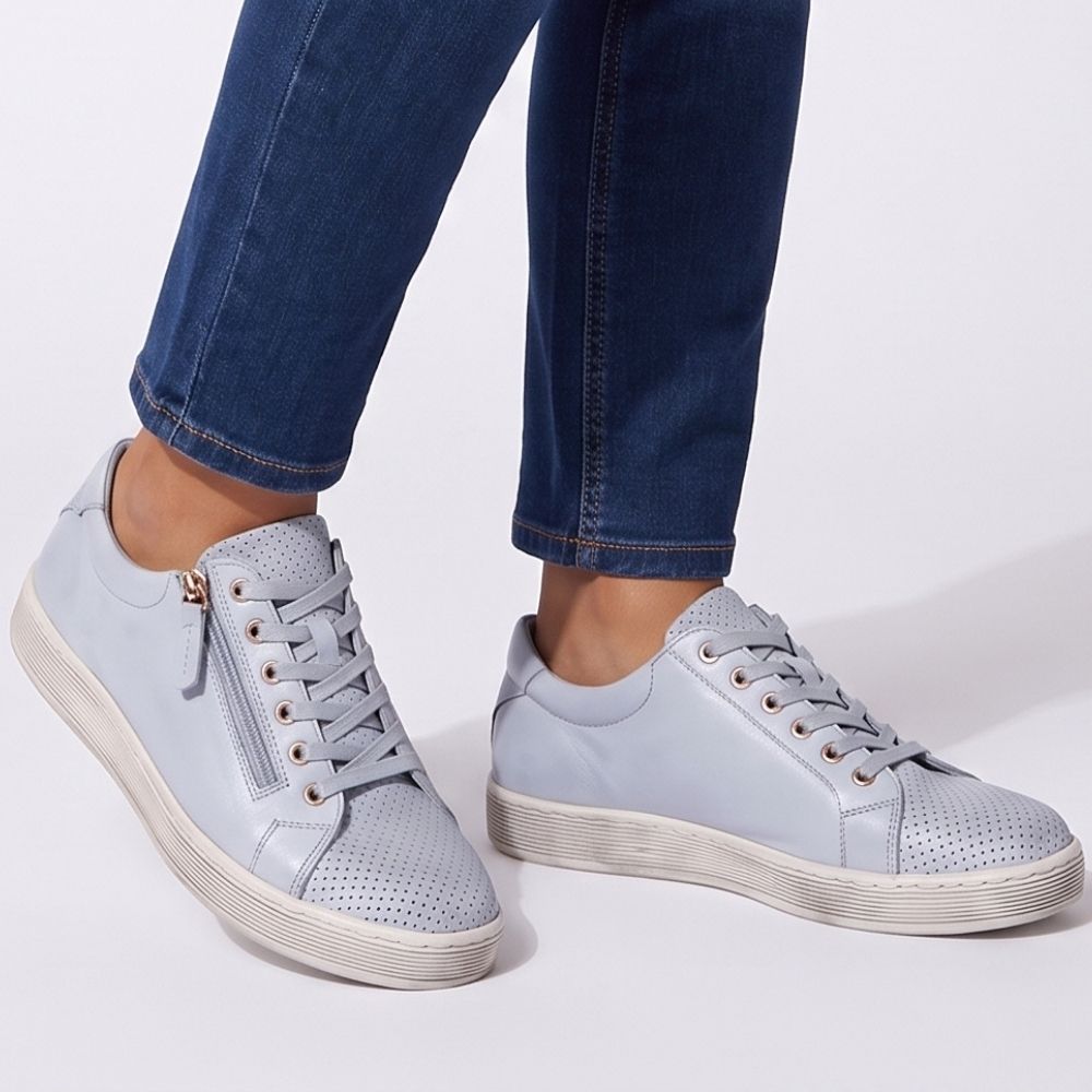 A person wearing dark blue jeans and a pair of pale blue lace-up sneakers with an outer zip entry and perforated details, standing against a studio background.