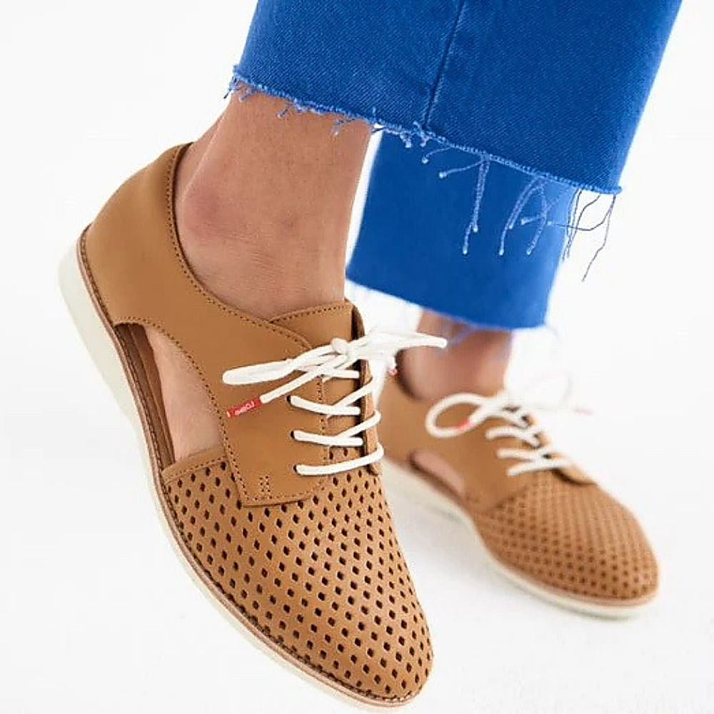 A person wearing blue jeans and a pair of tan leather lace-up shoes with a perforated toe cap, cut-out side panels and white soles, shown against a white studio background.
