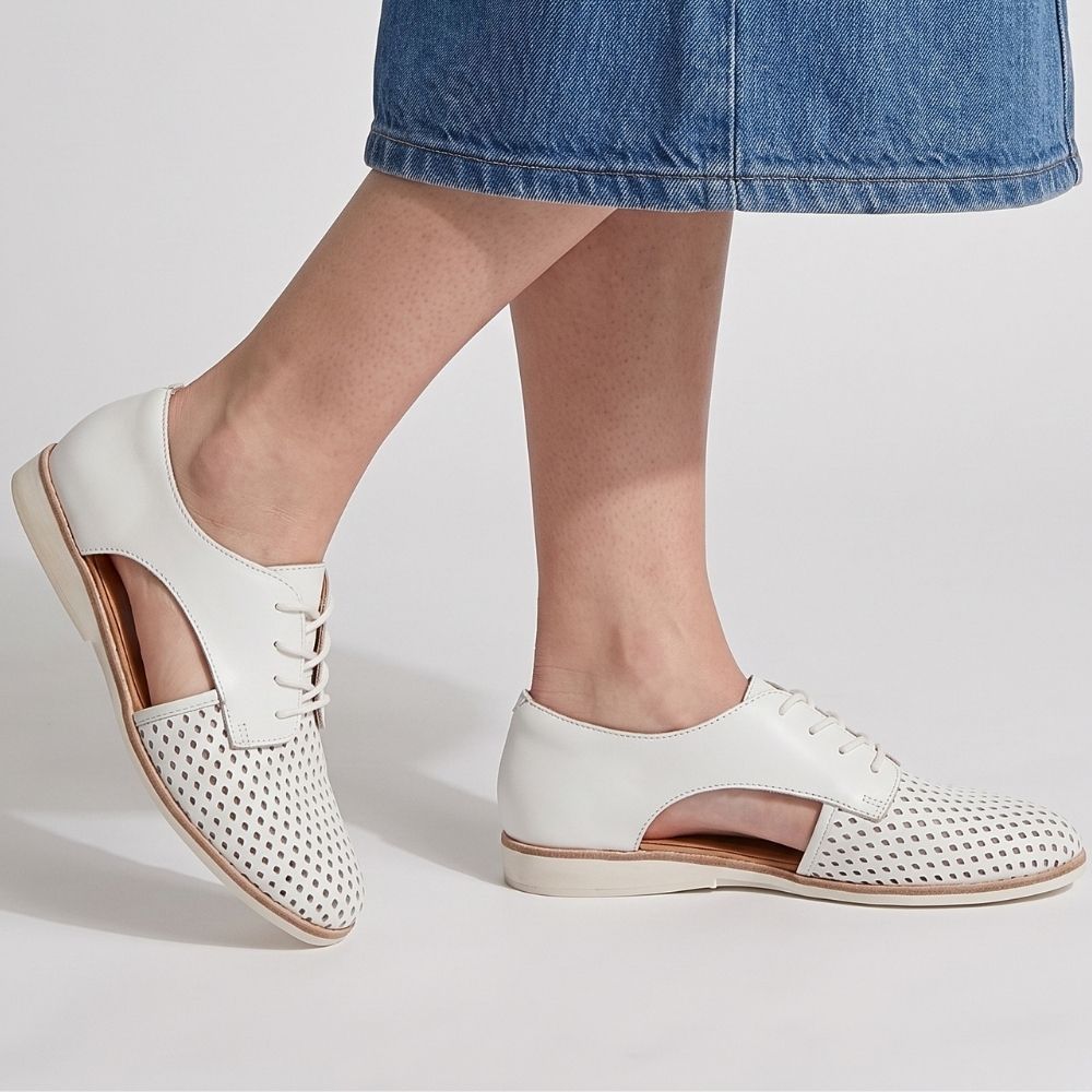 White leather lace-up shoes with perforated cutout detailing on the toe and cut-out side panels, shown on a model wearing blue denim shorts against a light gray studio background.