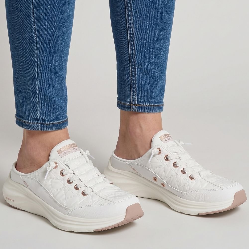 A person in blue jeans wearing a pair of white slip-on mule sneakers by SKECHERS featuring rose gold metallic details and a white sole, against a grey studio background.