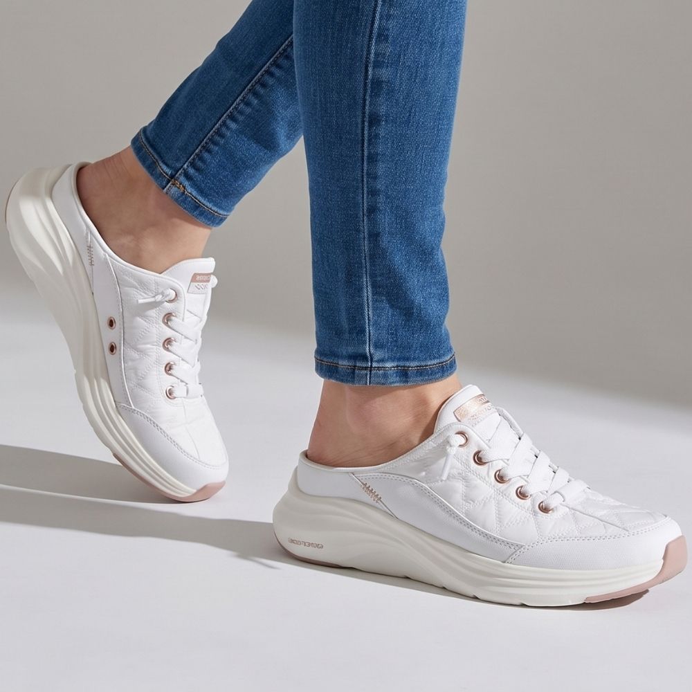 A person in blue jeans wearing a pair of white slip-on mule sneakers by SKECHERS featuring rose gold metallic details and a white sole, against a grey studio background.
