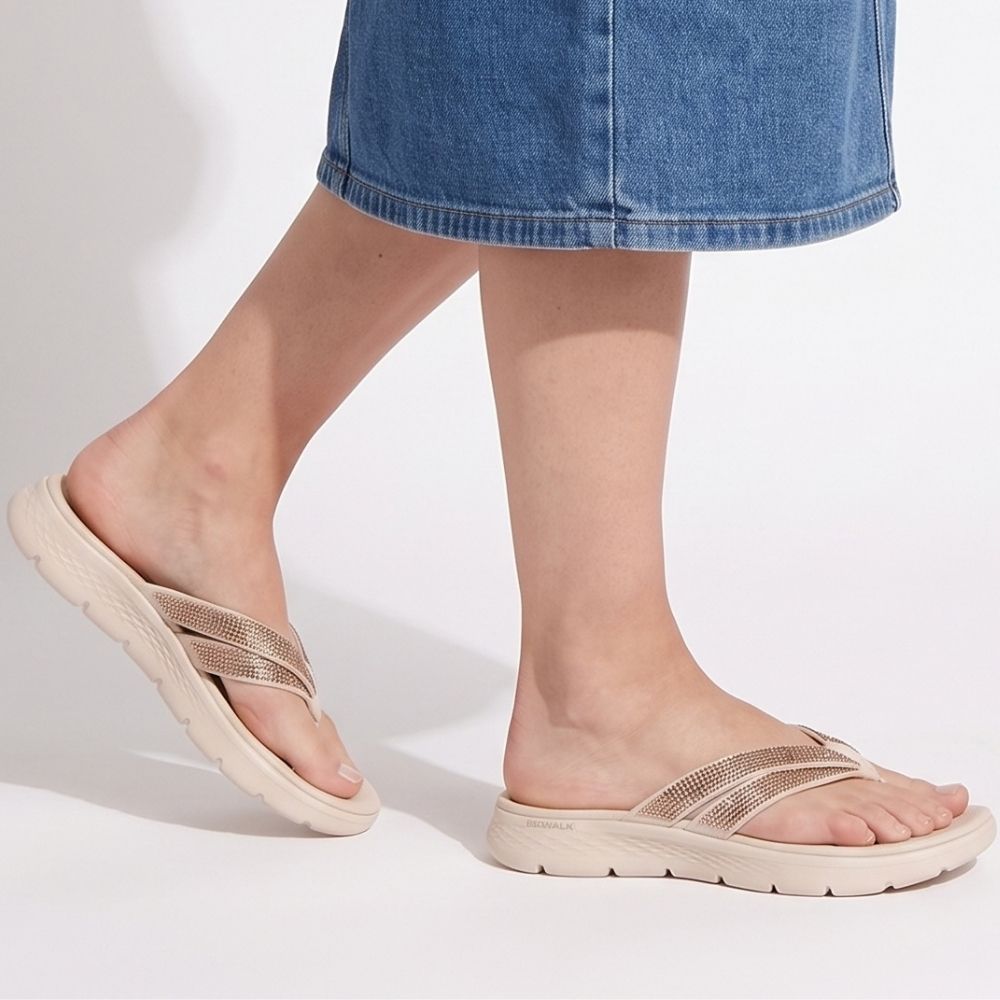 A person wearing beige slip on sandals with metallic rose-gold rhinestone embellished straps and a thick beige sole, paired with a blue denim skirt against a white studio background.