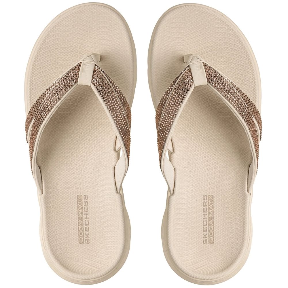 A top-down view of a pair of Skechers beige flip-flop sandals with metallic rose gold rhinestone embellished straps, on a white background. 