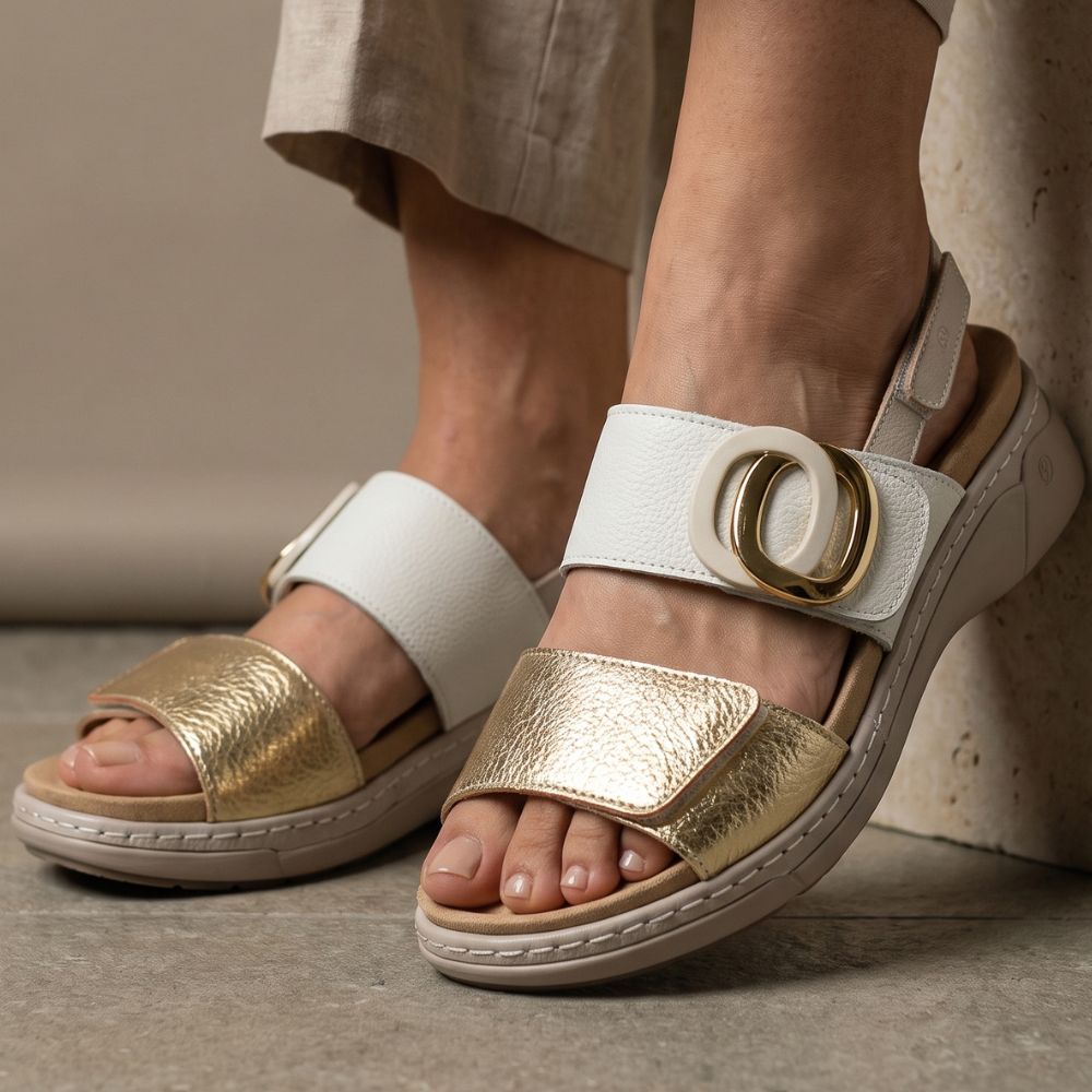 A close-up of a person wearing white and metallic gold platform sandals with wide straps and large circular gold buckle details, standing against a neutral stone-textured wall.