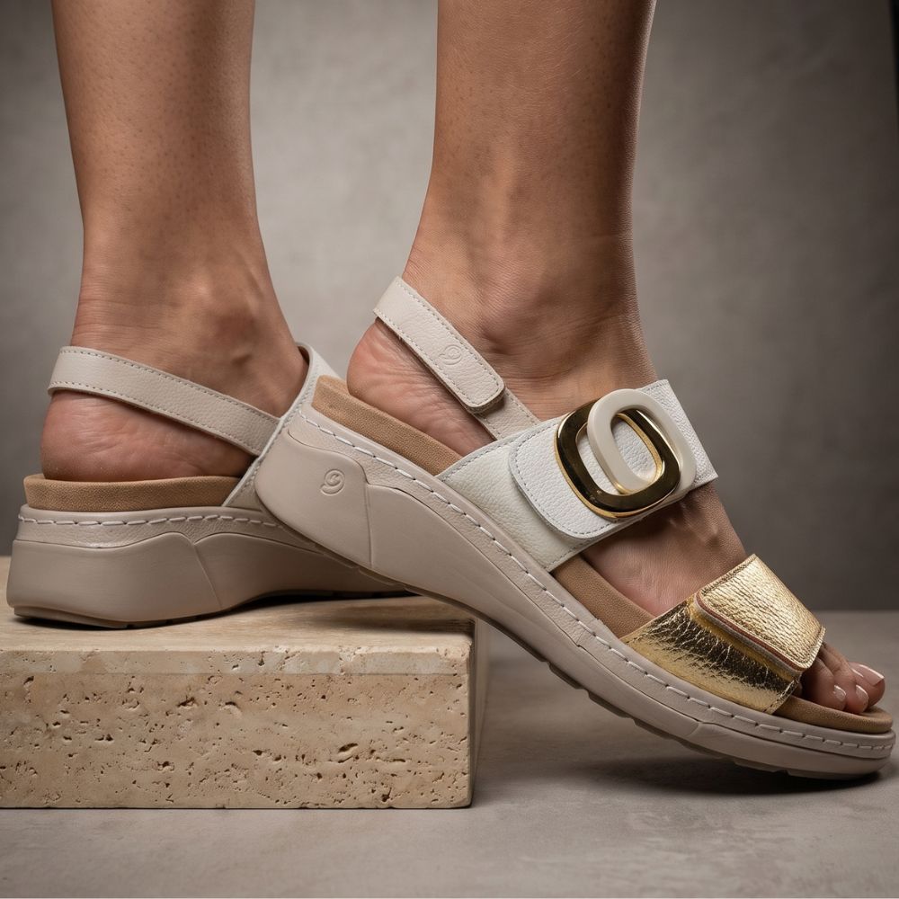A pair of women's platform sandals shown on feet, featuring a light beige faux-leather strap with a large gold buckle and a metallic gold toe strap, set against a neutral gray background.