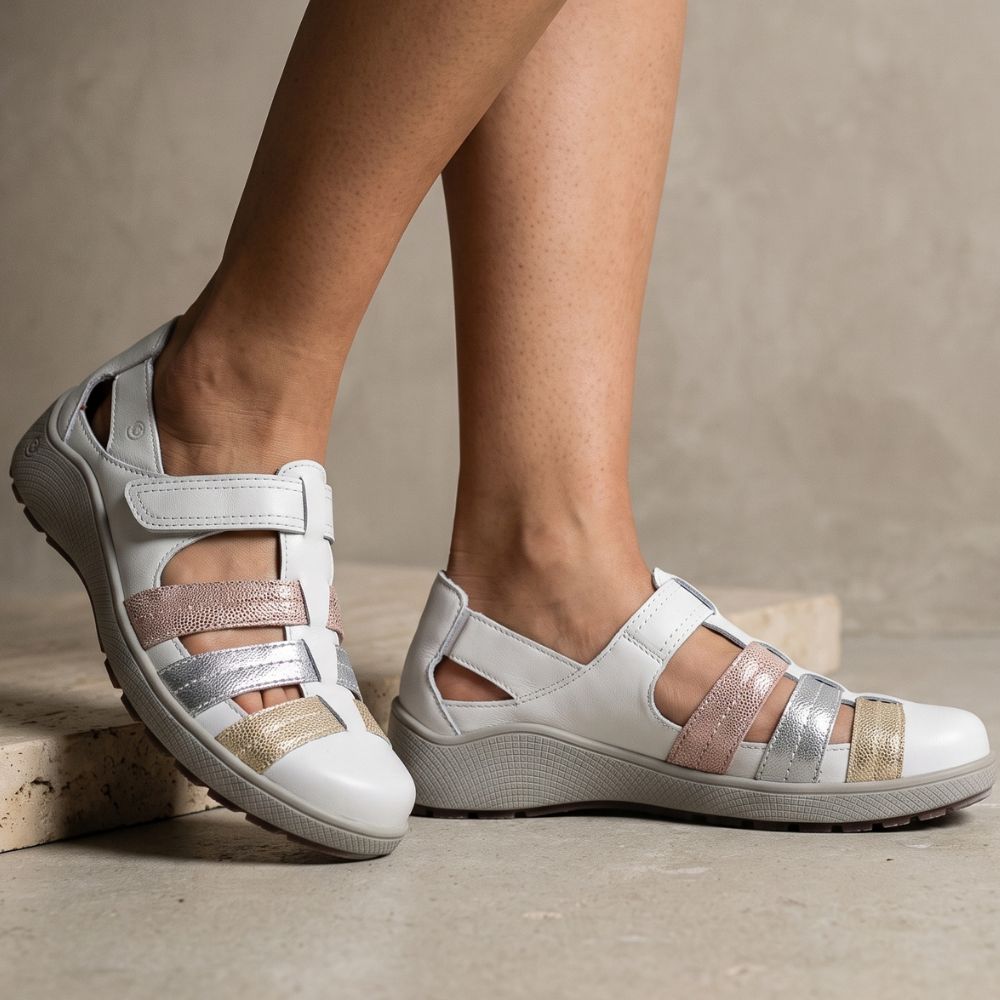 White sandals with metallic rose gold, silver, and gold straps, shown on a model's feet against a neutral concrete background.