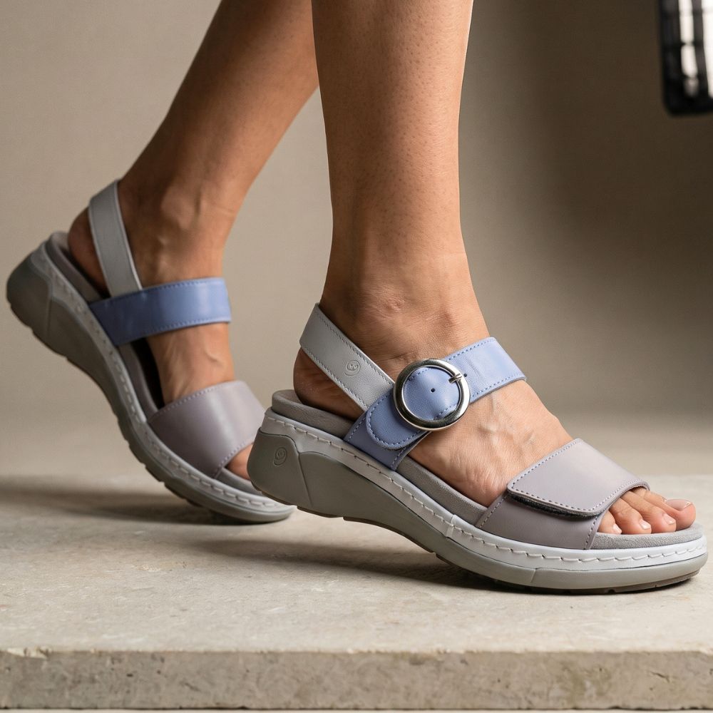 A pair of women's wedge sandals shown on a model's feet, featuring light gray and dusty blue straps with a silver-tone circular buckle, set against a neutral beige background.
