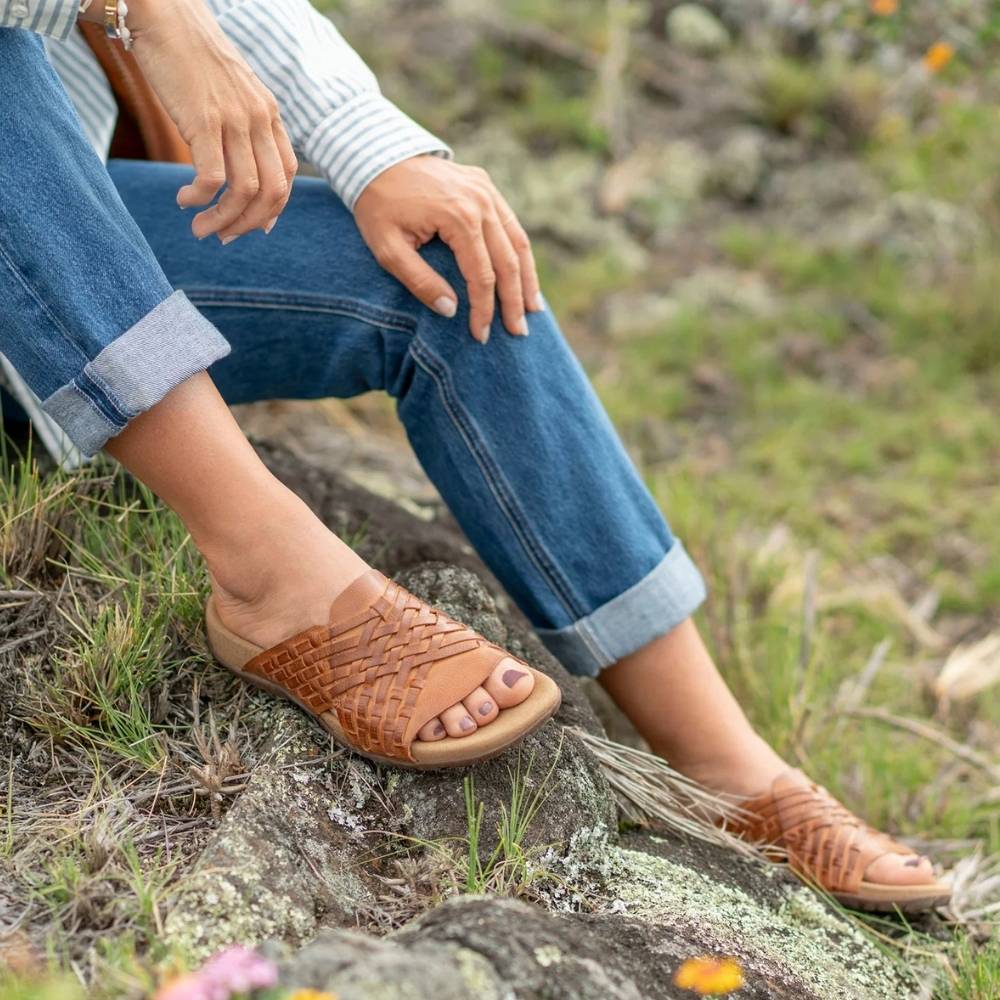 A person sitting outdoors on a rock wearing tan woven leather slide sandals with rolled-up blue jeans and a striped long-sleeve top.