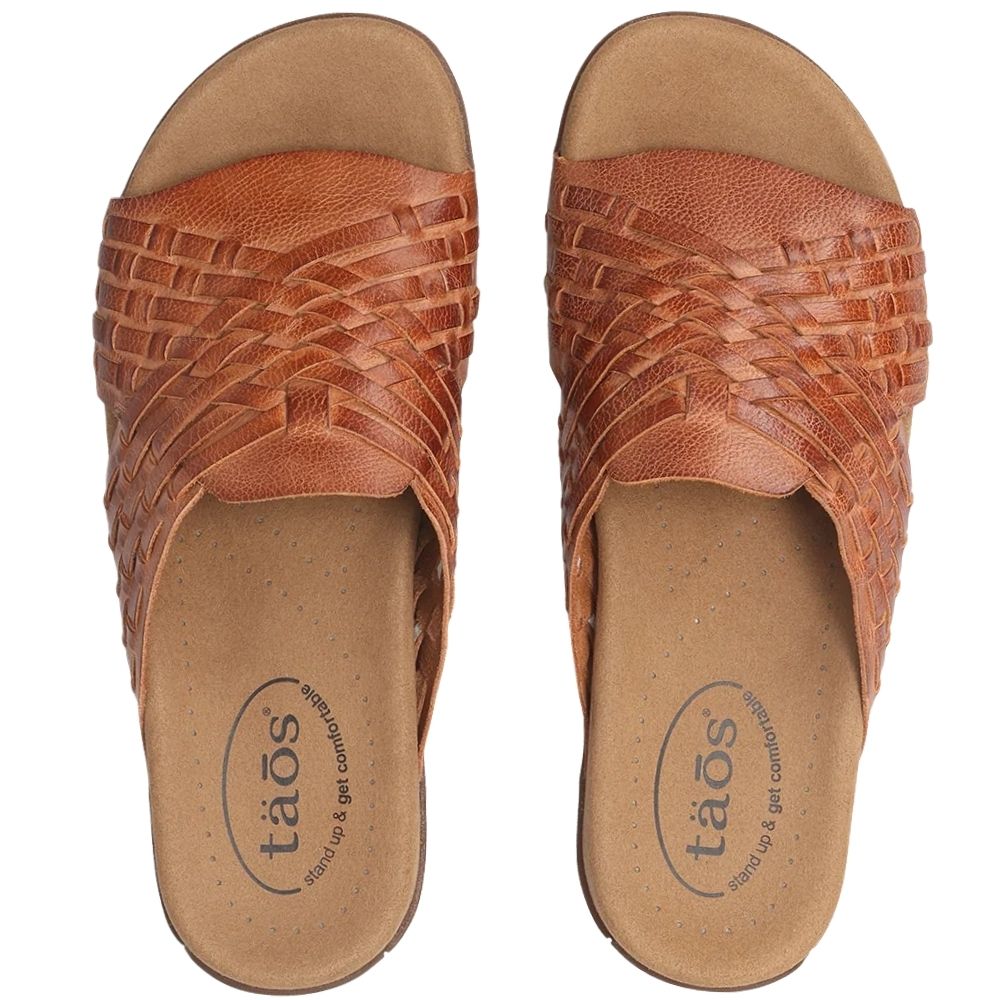 A pair of tan leather slide sandals with woven uppers, shown from a top-down view on a white background. The insole features the 'taos' brand logo.