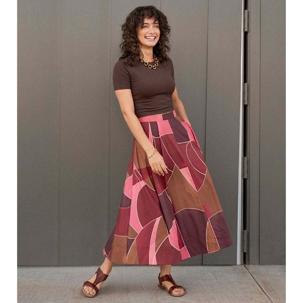 A woman modeling a brown short-sleeve top and a high-waisted midi skirt with an abstract geometric pattern in shades of pink, burgundy, and brown, standing against a gray paneled door with cognac leather T-strap sandals.