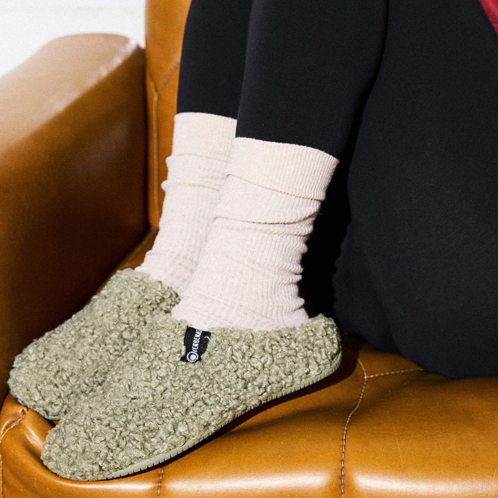 Person wearing white socks and green textured slippers sitting on a brown leather chair.
