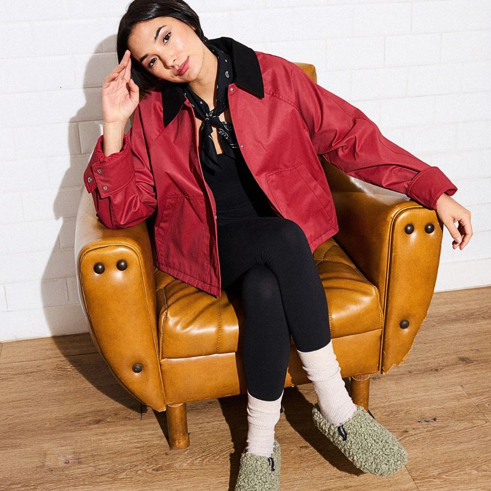 Woman wearing a red jacket sitting on a brown leather chair against a white brick wall wearing textured green slippers