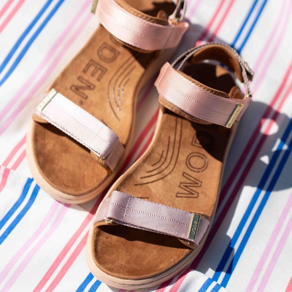 A pair of women's open-toe sandals with tan suede footbeds and woven blush, beige and tan adjustable straps, laid on a white fabric with pink and blue stripes.