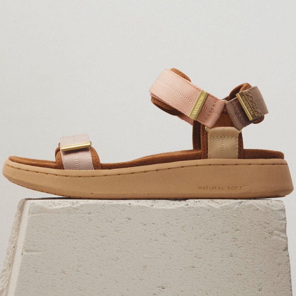 A single sandal from the brand Woden, shown in profile on a textured white block against a light gray background. The sandal features a tan sole, a woven blush toe strap with a gold detail, and a blush, tan and brown woven ankle strap with a gold metal detail.
