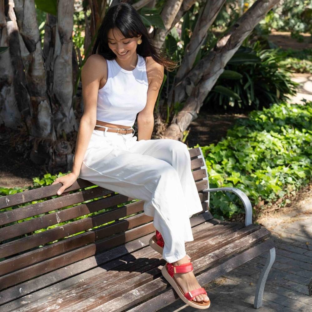 A woman sitting on a wooden park bench wearing a white sleeveless top and white wide-leg pants, with red sandals, in front of leafy green trees.