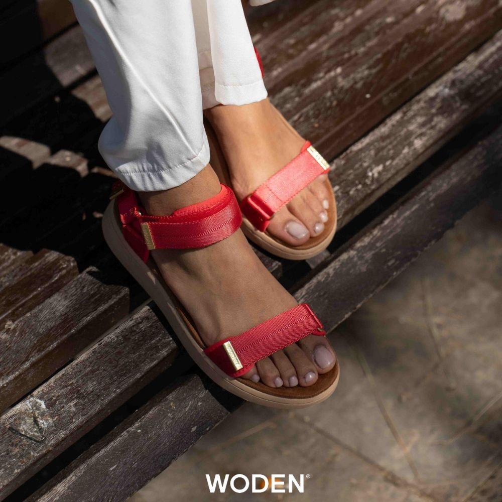 A person wearing red WODEN sandals with white pants, sitting on weathered wooden steps outdoors. The sandals feature red woven straps with small gold-tone metal accents and tan soles.