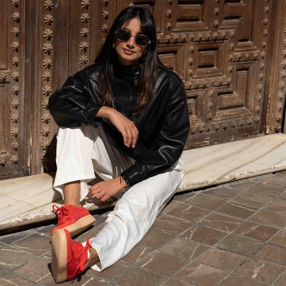 A woman sits on a stone ledge in front of a large wooden door, wearing a black faux-leather jacket, white wide-leg pants, and vibrant red sneakers.