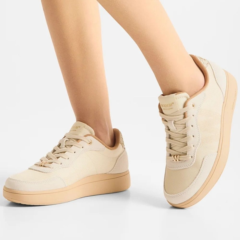A person wearing ivory sneakers with textured mesh, suede and leather panels with gold embellishments and beige soles, standing against a grey studio background.