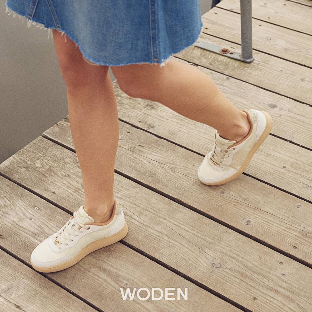 A person wearing ivory sneakers with textured mesh, suede and leather panels with gold embellishments and beige soles, walking on a boardwalk.